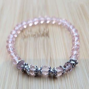 Op de afbeelding: Een lichtroze kralenarmband met zilverkleurige accenten. De armband heeft facetkralen en decoratieve metalen onderdelen. De kralen zijn geregen op een elastische band, waardoor een flexibel en stijlvol accessoire ontstaat.