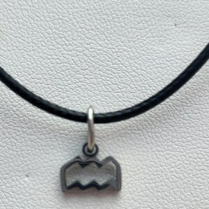 May include: A silver-colored pendant necklace featuring a mountain range design. The pendant hangs from a black cord. The necklace is displayed against a textured, off-white background.