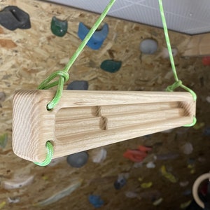 Puede incluir: Una presa de escalada de madera con tres agarres, suspendida del techo con una cuerda verde. La presa está hecha de madera clara y tiene un acabado natural.