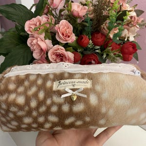 May include: A brown and white spotted pencil case with a white lace zipper and the text "princess-made". The case has a small white bow and a gold heart charm. A bouquet of pink and red flowers is in the background.