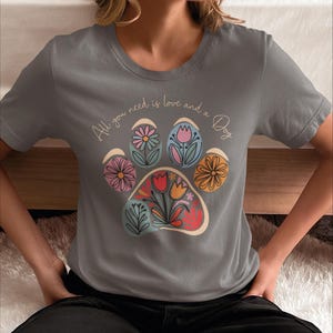 May include: Gray t-shirt with a paw print design made of colorful flowers. The text "All you need is love and a Dog" is written on the shirt.
