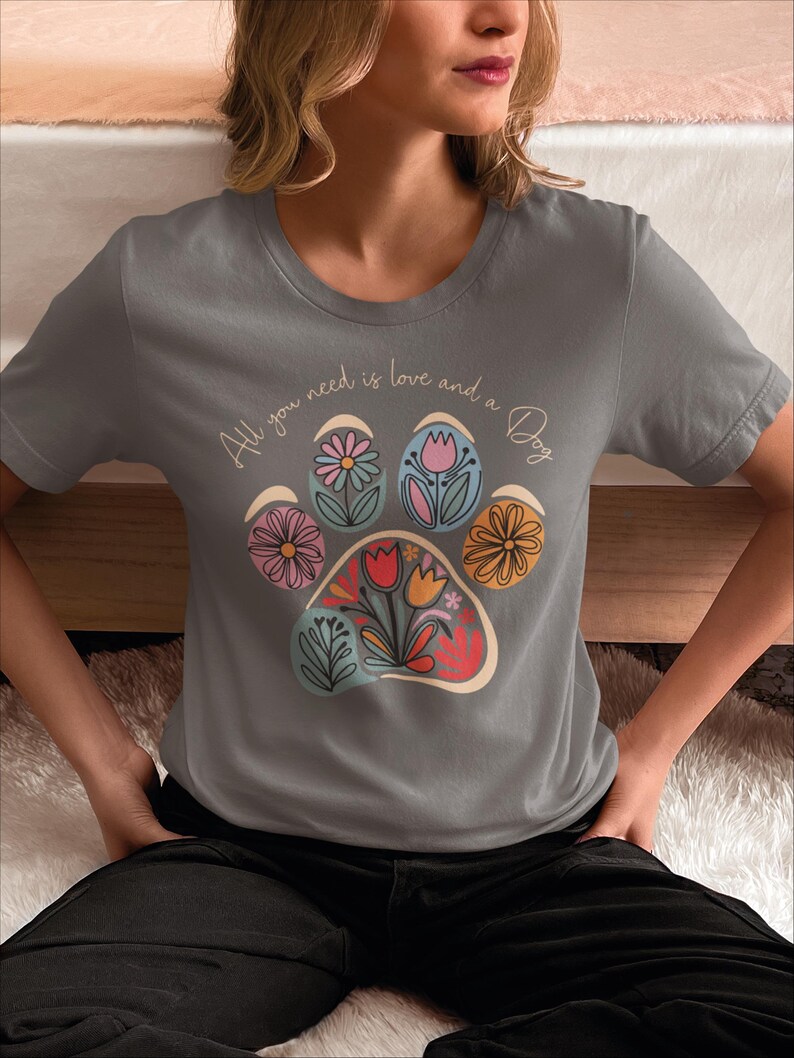 May include: Gray t-shirt with a paw print design made of colorful flowers. The text "All you need is love and a Dog" is written on the shirt.