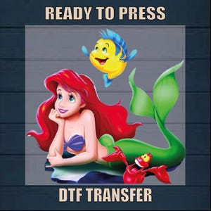 May include: A DTF transfer design featuring Ariel, the Little Mermaid, with her red hair, green tail, and a purple seashell bra. She is swimming with a yellow and blue fish. The design says "Ready to Press" and "DTF Transfer".