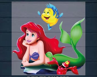 May include: A DTF transfer design featuring Ariel, the Little Mermaid, with her red hair, green tail, and a purple seashell bra. She is swimming with a yellow and blue fish. The design says "Ready to Press" and "DTF Transfer".