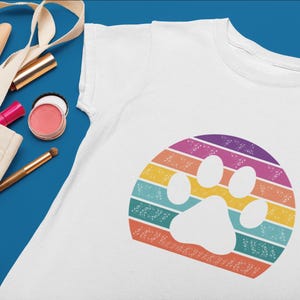 May include: White t-shirt with a rainbow paw print design. The paw print is in the center of the shirt and is surrounded by a rainbow of colors.
