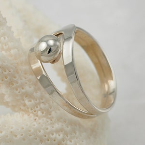 Puede incluir: Un anillo de plata con una cuenta redonda y lisa de plata en el centro. El anillo está hecho de dos bandas que se cruzan y envuelven la cuenta.