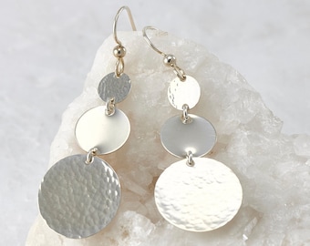 May include: Silver dangle earrings with three hammered circular discs hanging from a gold hook.