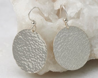 May include: A pair of silver earrings with a hammered, textured, circular design. The earrings are hanging from a white surface.