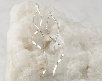 May include: A pair of silver dangle earrings with a twisted design. The earrings are displayed on a white rock.