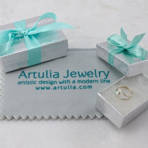 Puede incluir: Un anillo de plata con una piedra de &oacute;palo blanco en una peque&ntilde;a caja blanca con una cinta turquesa. La caja est&aacute; sobre un pa&ntilde;o de limpieza gris con el texto "Artullia Jewelry, artistic design with a modern line, www.artullia.com".
