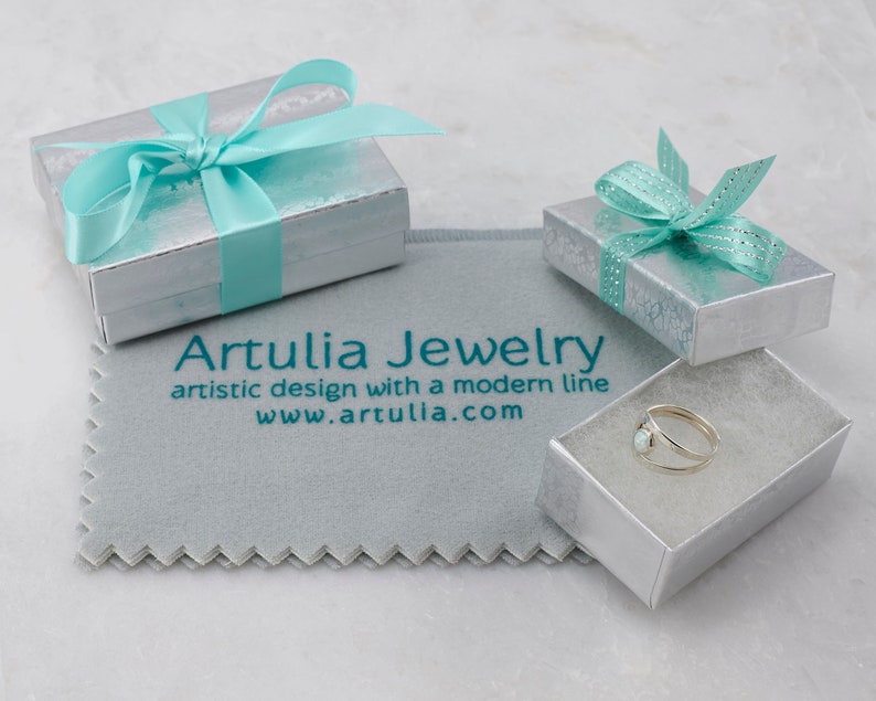 Puede incluir: Un anillo de plata con una piedra de &oacute;palo blanco en una peque&ntilde;a caja blanca con una cinta turquesa. La caja est&aacute; sobre un pa&ntilde;o de limpieza gris con el texto "Artullia Jewelry, artistic design with a modern line, www.artullia.com".
