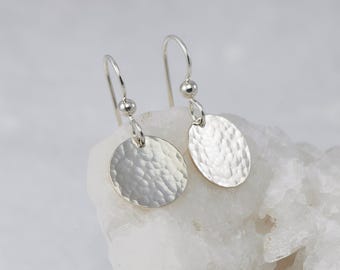 May include: A pair of silver drop earrings featuring circular discs with a hammered, textured finish. Each disc is suspended from a simple silver hook wire by a small, smooth silver bead, creating a reflective and metallic appearance.