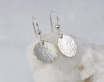 May include: A pair of silver dangle earrings with hammered round disc charms. The earrings have a simple, minimalist design.