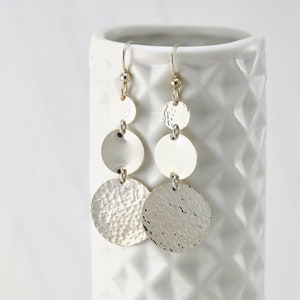 Puede incluir: Pendientes de plata con tres discos redondos, los dos inferiores son texturizados y el superior es liso. Los pendientes cuelgan de un gancho dorado.