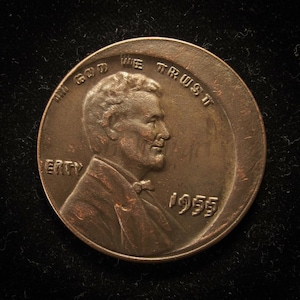 May include: A copper-colored 1955 Lincoln penny. The obverse features a profile of Abraham Lincoln, with the inscriptions "IN GOD WE TRUST" and "LIBERTY." The year "1955" is also visible.