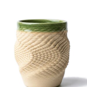 Pode incluir: Uma caneca de cerâmica bege com um padrão texturizado semelhante a tecido no corpo. A borda é de cor verde sólida. A caneca tem um formato arredondado e provavelmente é usada para bebidas.
