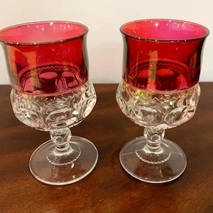 May include: Two matching stemmed goblets featuring a deep red glass upper bowl and a clear glass base. The lower portion of the bowl and the stem display a textured, circular pattern, while the clear circular foot provides stability for each glass.
