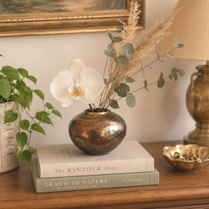 May include: A round, metallic bronze vase holds a single white orchid, dried pampas grass, and eucalyptus stems. The vase rests on two stacked books titled The Kinfolk Home and Drawn to Nature, positioned near a green plant and a gold bowl.