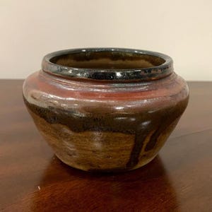 May include: A small, round ceramic bowl with a wide opening and a thick, dark rim. The exterior features a layered glaze with earthy tones of brown, tan, and a band of deep red, with dark brown glaze drips running down the sides of the vessel.