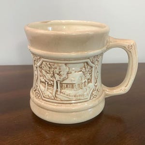 Peut inclure: Une tasse en céramique de couleur crème avec un motif en relief sur le devant. Le panneau central représente une maison entourée d'arbres dans un cadre octogonal. La poignée présente une forme géométrique avec des accents circulaires.