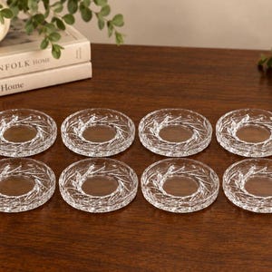 May include: A set of eight clear glass coasters arranged in two rows of four. Each circular coaster features a textured, radial leaf-like pattern etched into the glass, creating a decorative border around a smooth, transparent centre.