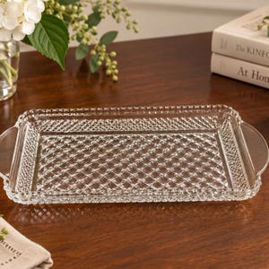 May include: A rectangular clear glass serving tray featuring a textured diamond-patterned base and a scalloped edge. The tray includes two smooth, integrated handles on opposite sides, providing a functional design for serving items.