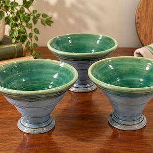 May include: Three ceramic footed bowls featuring a glossy teal interior and a matte light blue exterior. Each bowl has a wide, flared rim and a tapered base with visible horizontal ridges and dark brown accents along the edges and the footed base.