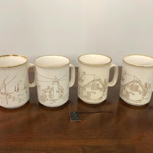 May include: A set of four off-white ceramic mugs with gold-toned rims, each featuring a brown line-art illustration of a coastal scene, including a lighthouse and houses. A small black rectangular tag with white text is attached to one mug.