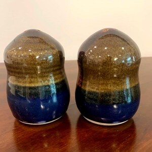May include: A pair of ceramic salt and pepper shakers with a rounded, bulbous shape. Each shaker features a tri-colour glaze transition, starting with a glossy brown top, a dark charcoal middle band, and a deep blue base with a white bottom rim.