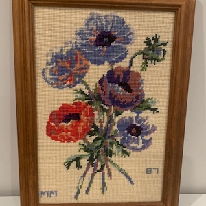 Può includere: Un ricamo a punto croce incorniciato raffigurante un bouquet di fiori nei toni del blu, viola e rosso su uno sfondo in tessuto color crema. La cornice rettangolare in legno ha una finitura marrone medio. Le lettere MIM e LB sono ricamate.