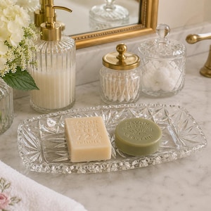 May include: A rectangular clear glass soap dish with a starburst pattern holds a rectangular beige soap bar embossed with Savon Extra Pur 72% and a round green soap bar embossed with Savon Extra Doux Verveine, set on a white marble surface.