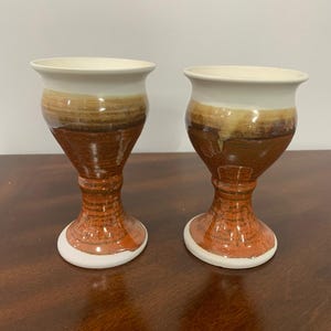 Peut inclure: Deux gobelets en céramique dotés d'un bord évasé et d'une base sur pied. La partie supérieure est de couleur crème, passant à un vernis brun chaud avec des coulures visibles. La tige inférieure et la base ont une finition en argile brun rougeâtre.