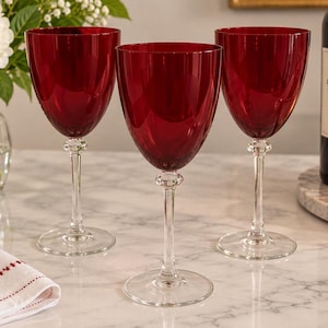 May include: Three wine glasses with deep red bowls and clear glass stems. Each glass features a rounded bowl shape, a slender stem with a decorative knob detail, and a circular base. The glasses are arranged in a row, showcasing the translucent red glass.