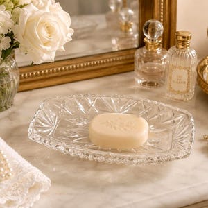 May include: An oval clear glass soap dish featuring a faceted geometric starburst pattern on the exterior surface. The dish holds a single smooth, light beige oval bar of soap with a subtle embossed floral design on its top surface.