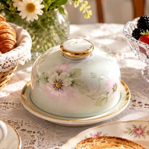 May include: A round porcelain butter dish with a dome-shaped lid featuring a pale green background, delicate pink and white floral patterns, and a gold-trimmed knob. The matching base plate also includes a thin gold rim along its circular edge.