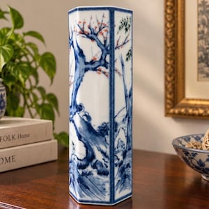 May include: A tall, hexagonal ceramic vase featuring a white base with intricate blue hand-painted illustrations of trees, branches, and foliage. The design includes subtle orange accents on the blossoms, with blue vertical borders on each edge.