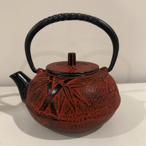 May include: A round cast iron teapot featuring a textured red finish with a black bamboo stalk relief pattern. The teapot includes a black lid with a central knob, a black curved spout, and a sturdy, arched black handle with a twisted rope texture.