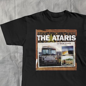 May include: Black t-shirt featuring the band name "THE ATARIS" above an image of a bus and two smaller photos. The text "In This Diary" is below the band name. The design is in a brown frame.