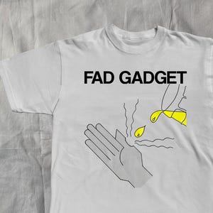 May include: A light gray t-shirt with the text "FAD GADGET" in black block letters. The shirt features a black and yellow graphic of a hand, a flame, and a dripping yellow liquid.