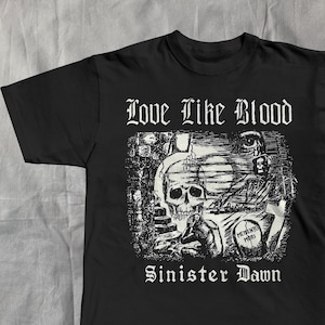 Op de afbeelding: Zwart T-shirt met een witte grafische afbeelding van een schedel, grafsteen en andere gotische elementen. De woorden "Love Like Blood" en "Sinister Dawn" staan boven en onder de afbeelding. Het shirt is gemaakt van een zacht materiaal.