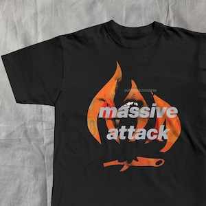 May include: Black t-shirt featuring the text "massive attack" in a white and orange flame design. Below the text is an orange knife graphic. The shirt is made of a soft material.