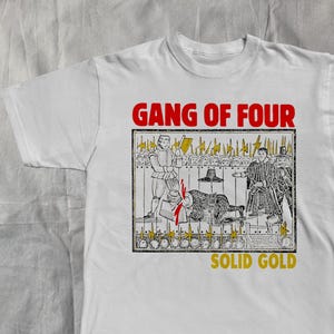 以下が含まれることがあります： 赤色の「GANG OF FOUR」の文字がプリントされた白いTシャツ。その下に、黒と白のイラストと黄色のアクセントがあります。イラストは歴史的な人物を描いており、下には黄色の「SOLID GOLD」の文字があります。