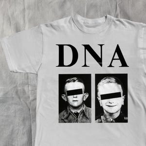 May include: White t-shirt with the word "DNA" printed in large, black capital letters. Below the text are two black and white images of children with their eyes obscured by black bars.