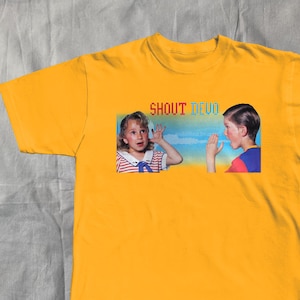 May include: Mustard yellow t-shirt with a graphic of two children gesturing, and the text "SHOUT DEVO" in a rainbow font. The children are set against a blue sky background. The shirt has a classic crew neck.