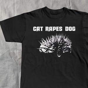 May include: Black t-shirt with the text "CAT RAPES DOG" in white block letters above a white graphic of a spiky, wheeled vehicle. The shirt is a crew neck style.