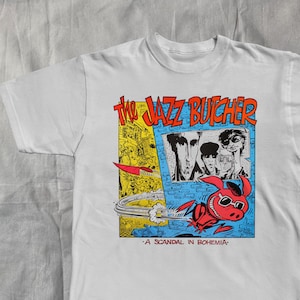 May include: White t-shirt featuring a colorful graphic design. The design includes the text "The Jazz Butcher" in orange, a band of people, and a red cartoon character with sunglasses. The text "A Scandal in Bohemia" is also present.