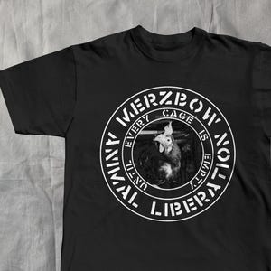 Może przedstawiać: Czarna koszulka z białym okrągłym nadrukiem. Grafika przedstawia koguta i tekst "MERZBOW" na górze, "EVERY CAGE IS EMPTY" pośrodku i "ANIMAL LIBERATION UNTIL" na dole. Koszulka wykonana jest z miękkiego materiału.