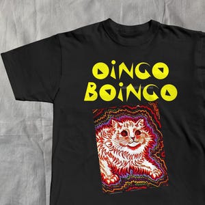 Pode incluir: T-shirt preto com a inscrição amarela "Dingo Boingo" acima de uma ilustração colorida de um gato. A ilustração do gato é vermelha e branca com uma borda psicodélica.