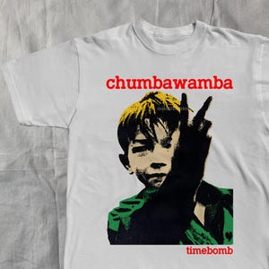 Puede incluir: Camiseta blanca con un gráfico de una persona con pelo amarillo y un brazo negro haciendo un gesto con la mano. El texto rojo "chumbawamba" está encima de la imagen, y la palabra "timebomb" está debajo.