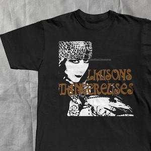 May include: Black t-shirt featuring a vintage-style graphic of a woman's face in white and brown, with the words "Liaisons Dangereuses" in brown. The design has a retro aesthetic, suitable for fans of vintage fashion and music.